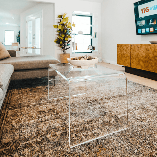 A clear acrylic waterfall edge coffee table in a living room in front of a couch decorated with a stone bowl containing potpourri.