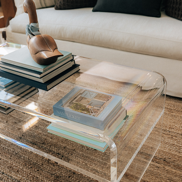 Clear acrylic coffee table with 1 interior shelf displaying books and decorative items in a living room setting.