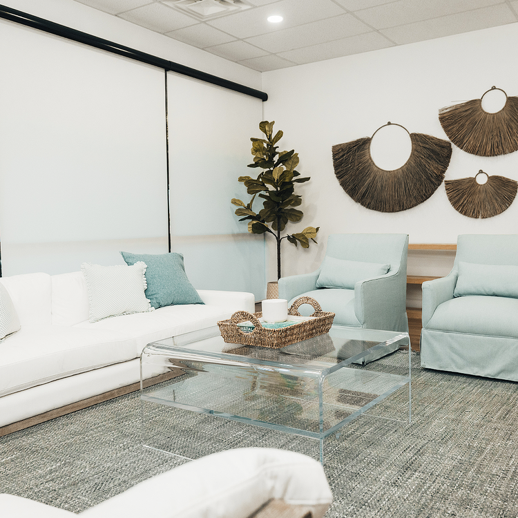 A clear acrylic square coffee table with a waterfall edge design and 1 interior shelf, placed in a living room setting with a white sofa, light blue easy chairs and decorative items.