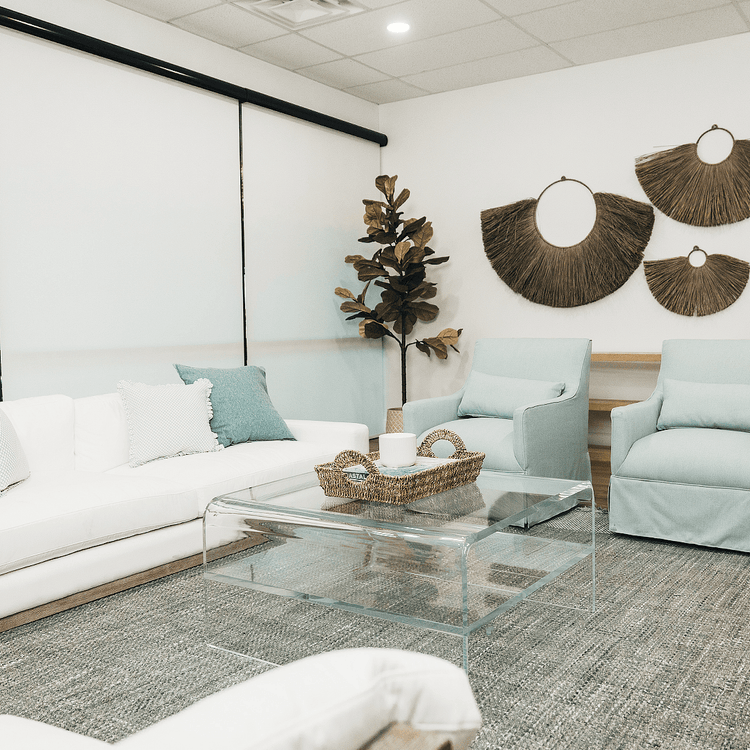 A clear acrylic square coffee table with a waterfall edge design and 1 interior shelf, placed in a living room setting with a white sofa, light blue easy chairs and decorative items.