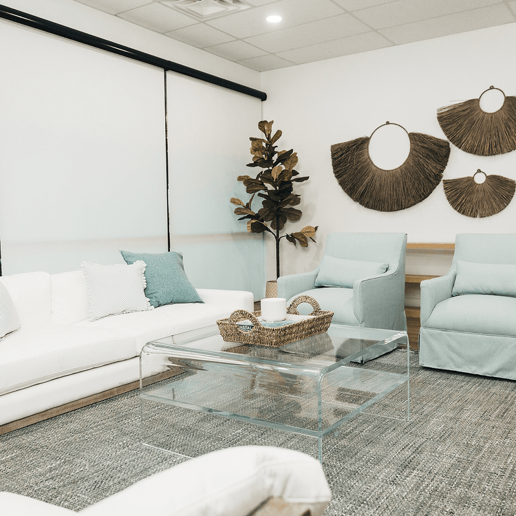 A clear acrylic waterfall edge coffee table in a living room setting in front of a couch featuring 1 interior shelf decorated with a basket and candle.