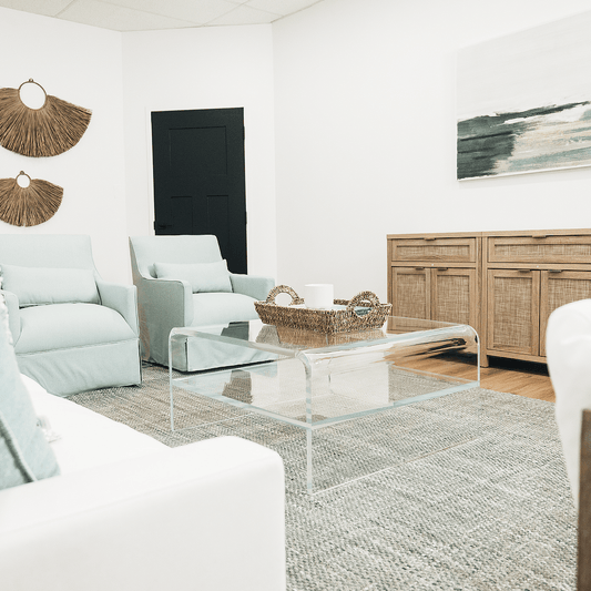 A clear acrylic square coffee table with a waterfall edge design and 1 interior shelf, placed in a living room setting with a white sofa, light blue easy chairs and decorative items.