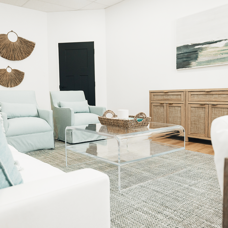 A clear acrylic square coffee table with a waterfall edge design and 1 interior shelf, placed in a living room setting with a white sofa, light blue easy chairs and decorative items.