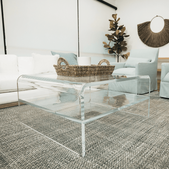 A clear acrylic waterfall edge coffee table in front of a couch featuring 1 interior shelf decorated with a basket and candle.