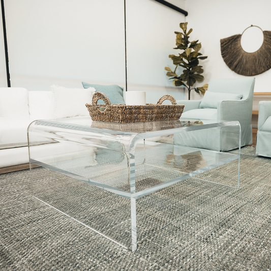 A clear acrylic square coffee table with a waterfall edge design and 1 interior shelf, placed in a living room setting with a white sofa, light blue easy chairs and decorative items.
