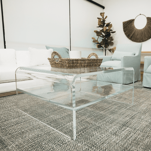 A clear acrylic square coffee table with a waterfall edge design and 1 interior shelf, placed in a living room setting with a white sofa, light blue easy chairs and decorative items.