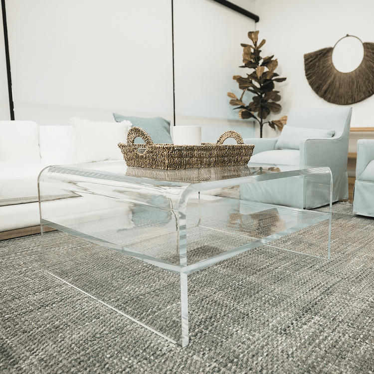 A clear acrylic square coffee table with a waterfall edge design and 1 interior shelf, placed in a living room setting with a white sofa, light blue easy chairs and decorative items.