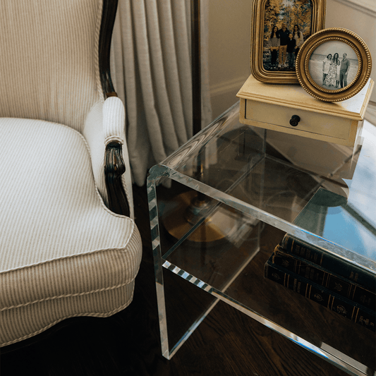 Clear acrylic side table with books and decorative items next to a white armchair in a room with a lamp and window.