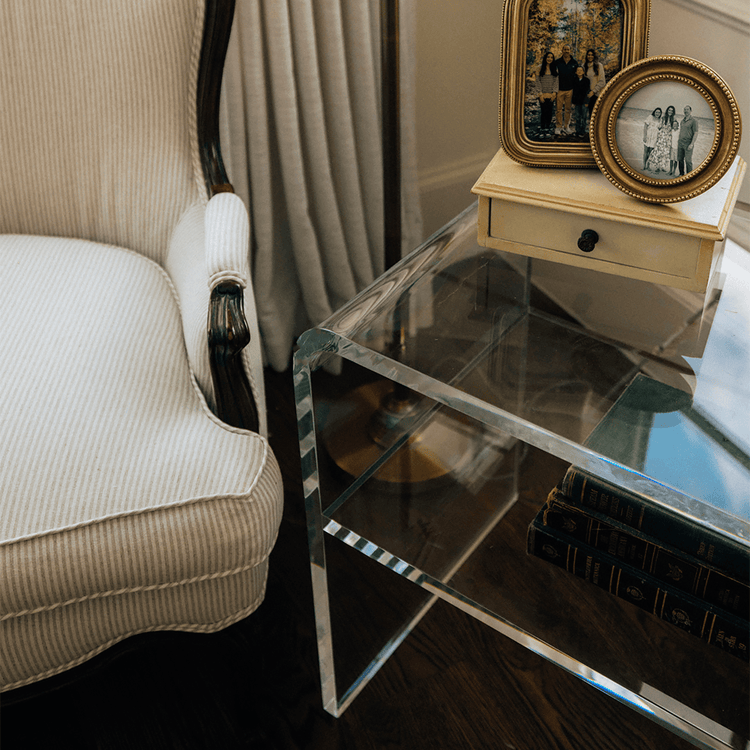 Clear acrylic side table with books and decorative items next to a white armchair in a room with a lamp and window.