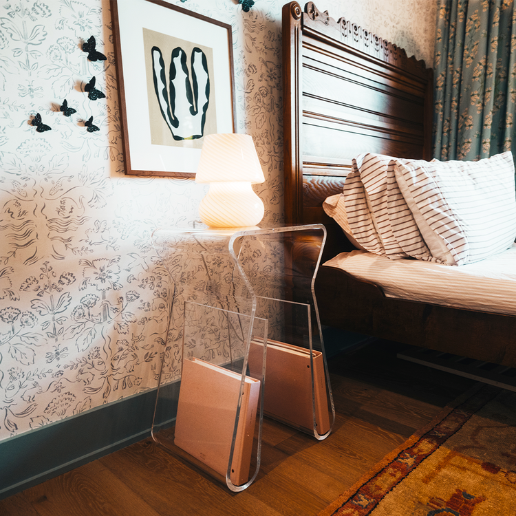 An acrylic lucite side table featuring legs that curve up into the inside of the table to double as a book or magazine rack displaying a lamp sitting beside a bed doubling as a nightstand.