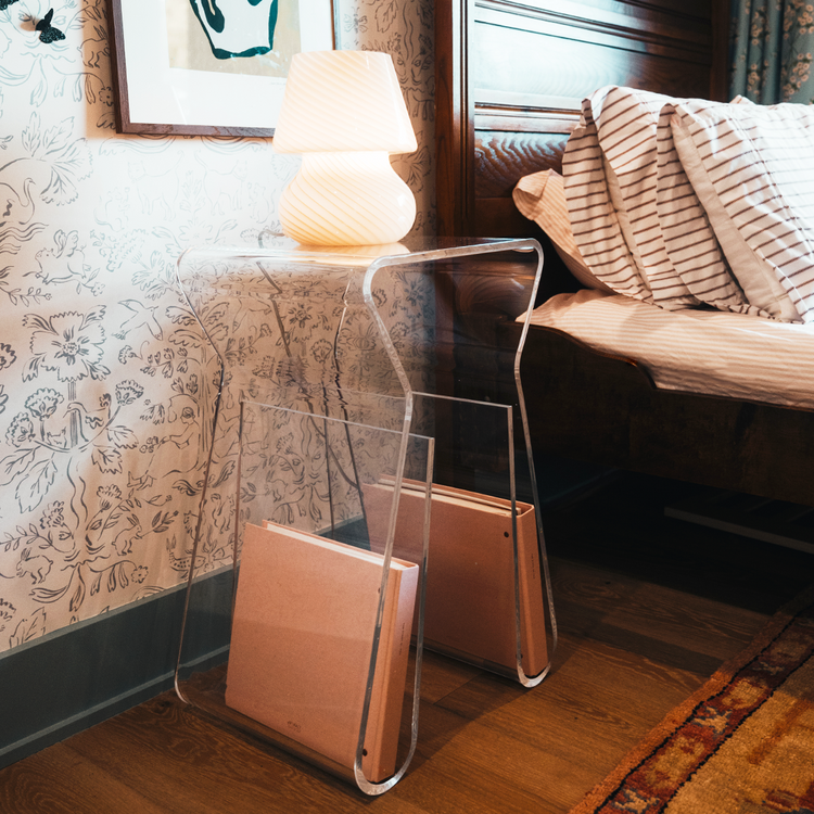 An acrylic lucite side table featuring legs that curve up into the inside of the table to double as a book or magazine rack displaying a lamp sitting beside a bed doubling as a nightstand.