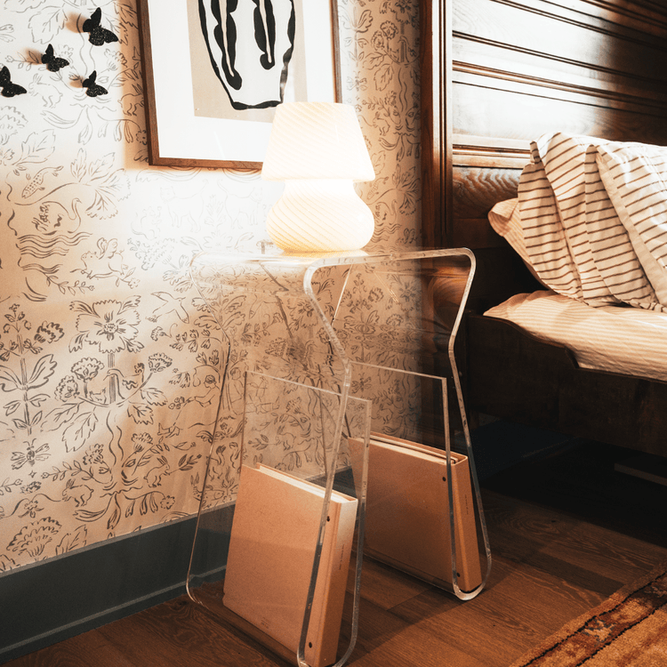 An acrylic lucite side table featuring legs that curve up into the inside of the table to double as a book or magazine rack displaying a lamp sitting beside a bed doubling as a nightstand.
