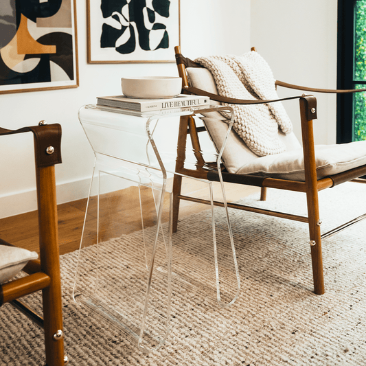 An acrylic lucite side table featuring legs that curve up into the inside of the table to double as a book or magazine rack sitting between 2 chairs in a living room.