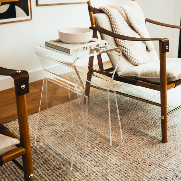 An acrylic lucite side table featuring legs that curve up into the inside of the table to double as a book or magazine rack sitting between 2 chairs in a living room.