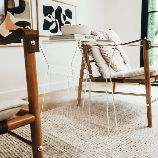 An acrylic lucite side table featuring legs that curve up into the inside of the table to double as a book or magazine rack sitting between 2 chairs in a living room.