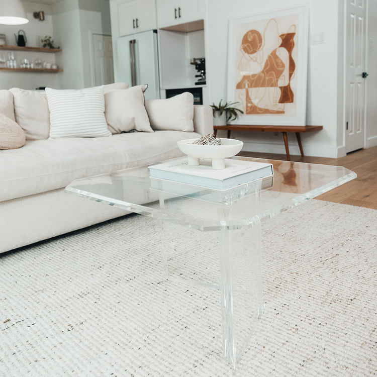 A clear acrylic lucite coffee table that features a thick u-shaped base displaying books and decor in a living room in front of a couch.