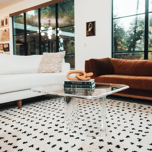A clear acrylic lucite coffee table that features a thick u-shaped base displaying books and decor in a living room in front of a couch.