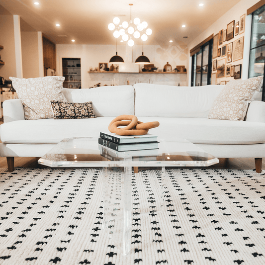 A clear acrylic lucite coffee table that features a thick u-shaped base displaying books and decor in a living room in front of a couch.