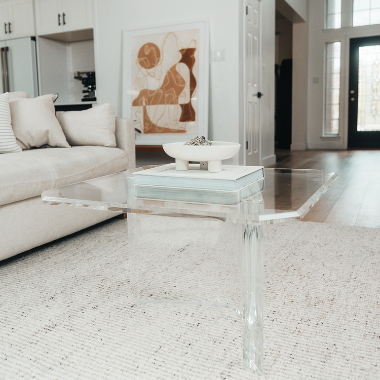 A clear acrylic lucite coffee table that features a thick u-shaped base displaying books and decor in a living room in front of a couch.