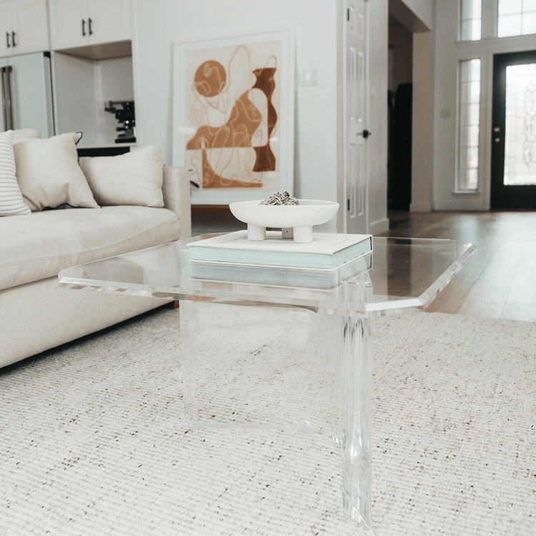 A clear acrylic lucite coffee table that features a thick u-shaped base displaying books and decor in a living room in front of a couch.
