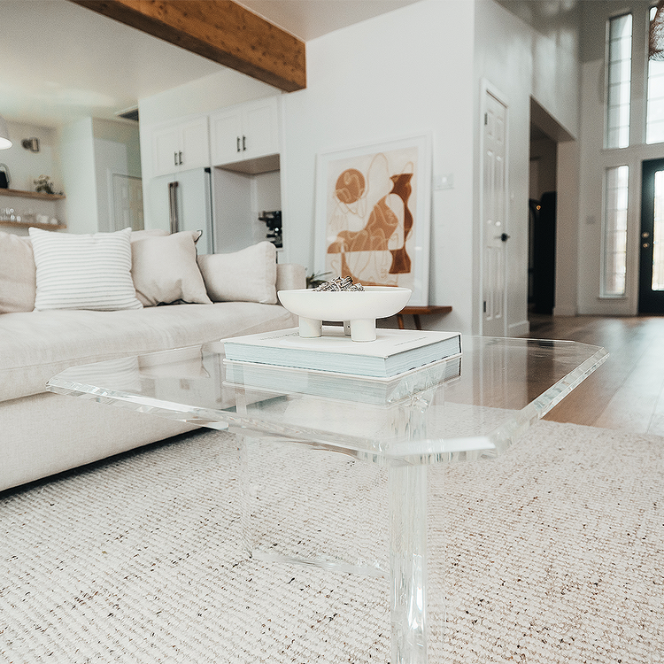 A clear acrylic lucite coffee table that features a thick u-shaped base displaying books and decor in a living room in front of a couch.