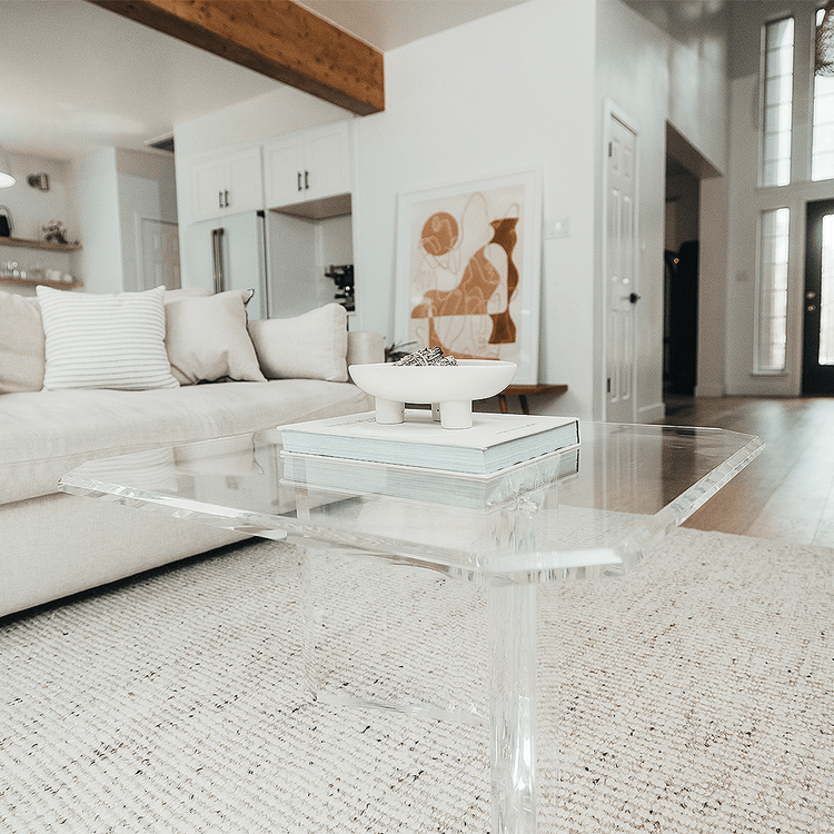 A clear acrylic lucite coffee table that features a thick u-shaped base displaying books and decor in a living room in front of a couch.