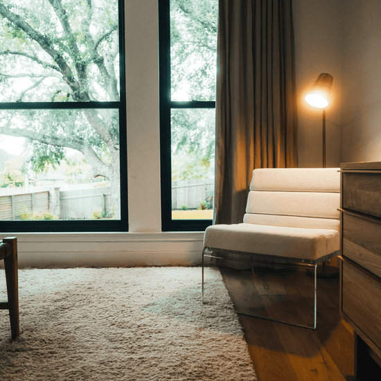 An acrylic lounge chair with a reading lamp behind, placed in the corner of a bedroom next to a dresser and picture window with curtains.