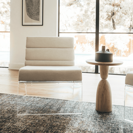 A clear acrylic lucite lounge chair with a white fabric seat and backrest, placed in a living room setting in front of large picture windows.