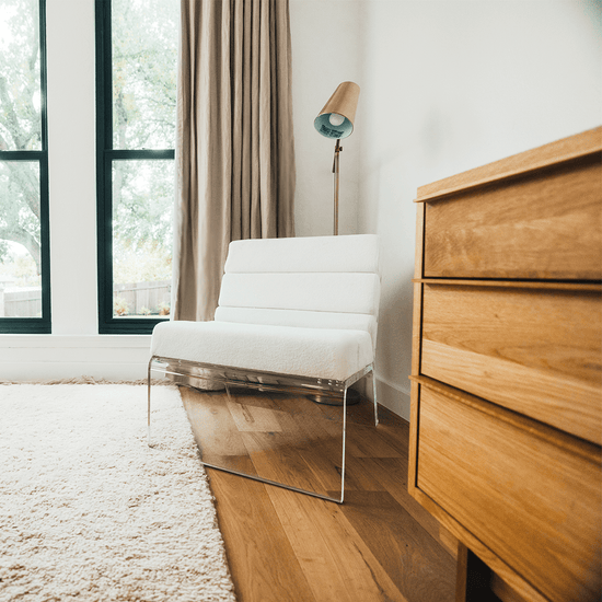 An acrylic lounge chair with a reading lamp behind, placed in the corner of a bedroom next to a dresser and picture window with curtains.