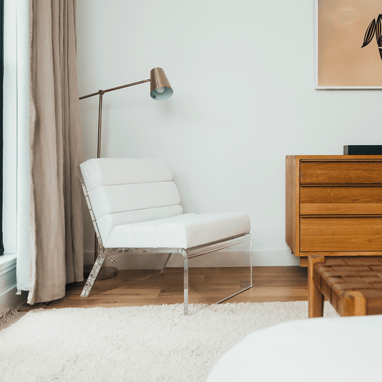 An acrylic lounge chair with a reading lamp behind, placed in the corner of a bedroom next to a dresser and picture window with curtains.
