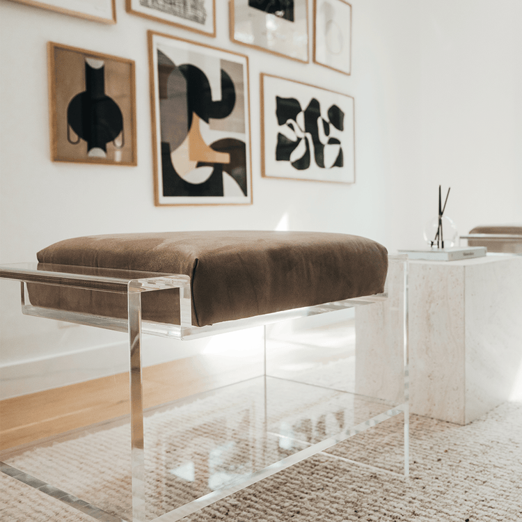 Closeup view of 1 of the two clear acrylic benches with brown cushions and storage shelf underneath, placed as extra seating in a modern living room with abstract art on the walls.