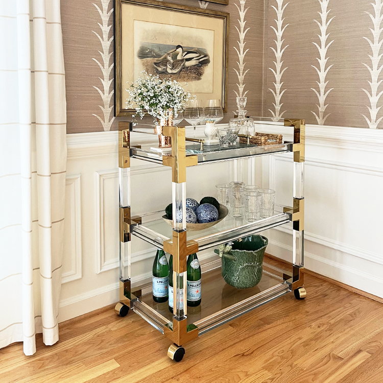 An acrylic lucite bar cart placed in a dining room corner featuring gold hardware, 3 shelves, and wheels for easy movement. The cart is decorated with flowers, spirts, glasses, and various bar accessories.