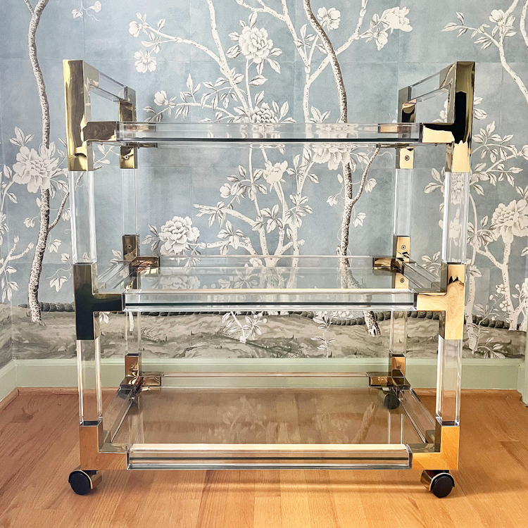 An acrylic lucite bar cart featuring gold hardware, 3 empty shelves, and wheels for easy movement.