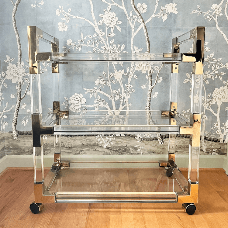 An acrylic lucite bar cart featuring gold hardware, 3 empty shelves, and wheels for easy movement.