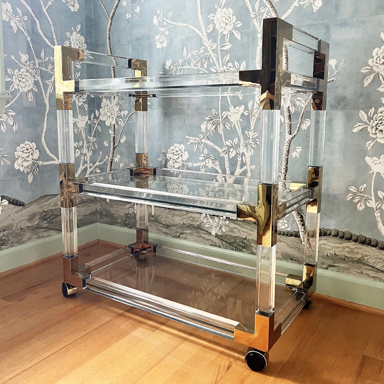 An acrylic lucite bar cart featuring gold hardware, 3 empty shelves, and wheels for easy movement.