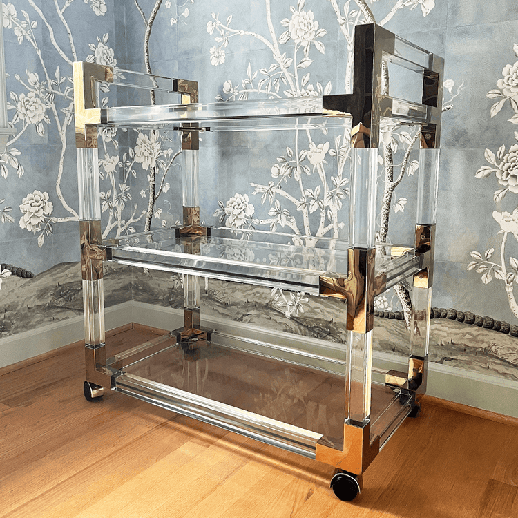 An acrylic lucite bar cart featuring gold hardware, 3 empty shelves, and wheels for easy movement.