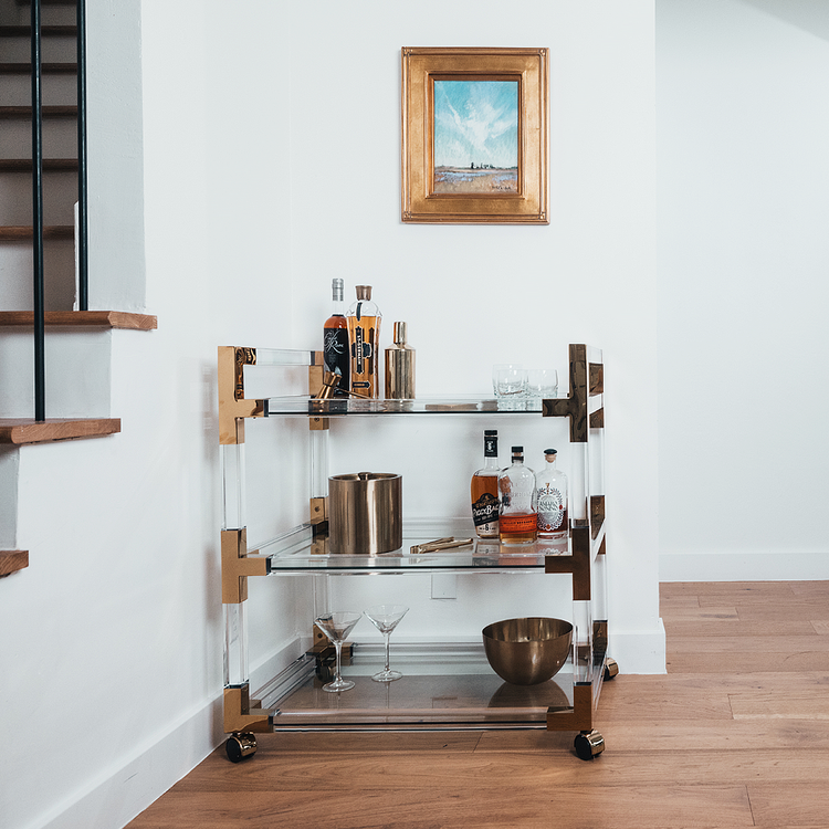An acrylic lucite bar cart placed in a dining room corner featuring gold hardware, 3 shelves, and wheels for easy movement. The cart is decorated with spirts, glasses, and various bar accessories.