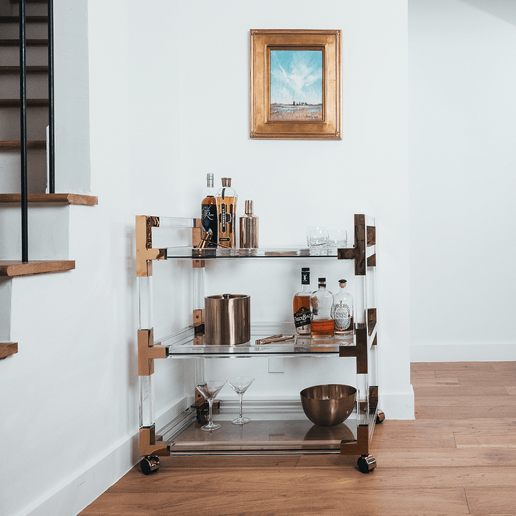 An acrylic lucite bar cart placed in a dining room corner featuring gold hardware, 3 shelves, and wheels for easy movement. The cart is decorated with spirts, glasses, and various bar accessories.
