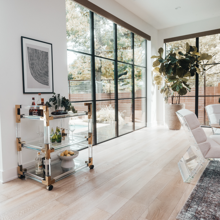 An acrylic lucite bar cart placed in a living room next to windows featuring gold hardware, 3 shelves, and wheels for easy movement. The cart is decorated with a plant, spirts, glasses, and various bar accessories.