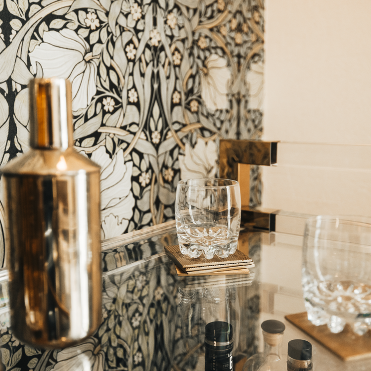 A closeup of the top shelf of an acrylic lucite bar cart featuring gold hardware displaying spirts, glasses and various bar accessories.