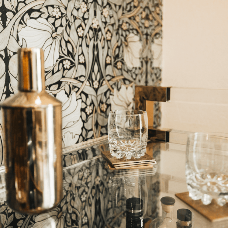 A closeup of the top shelf of an acrylic lucite bar cart featuring gold hardware displaying spirts, glasses and various bar accessories.