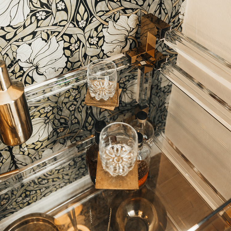 A closeup of the top shelf of an acrylic lucite bar cart featuring gold hardware displaying spirts, glasses and various bar accessories.