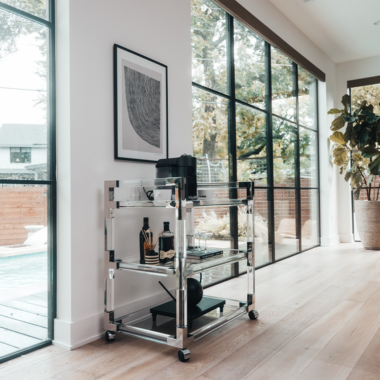 An acrylic lucite bar cart placed in a living room featuring silver hardware, 3 shelves, and wheels for easy movement. This cart is also functioning as a coffee bar displaying a coffee pot, cups, spirts, and glasses.