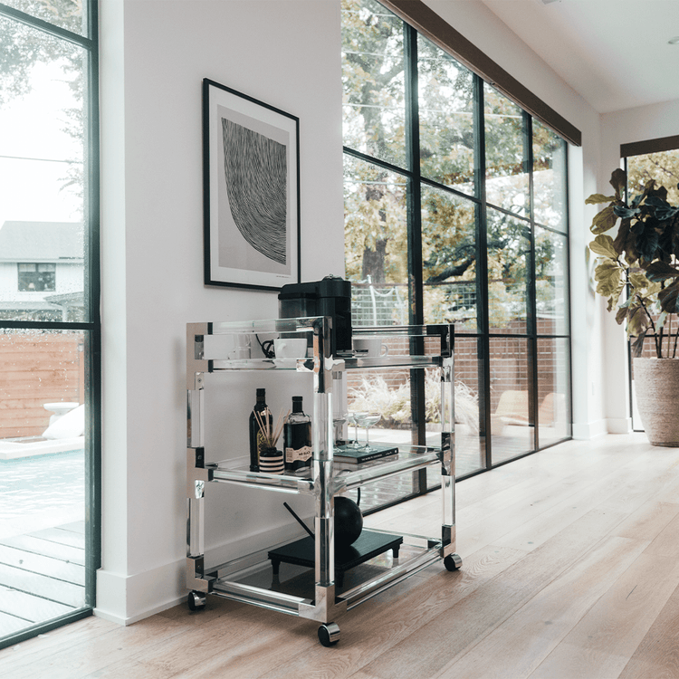 An acrylic lucite bar cart placed in a living room featuring silver hardware, 3 shelves, and wheels for easy movement. This cart is also functioning as a coffee bar displaying a coffee pot, cups, spirts, and glasses.