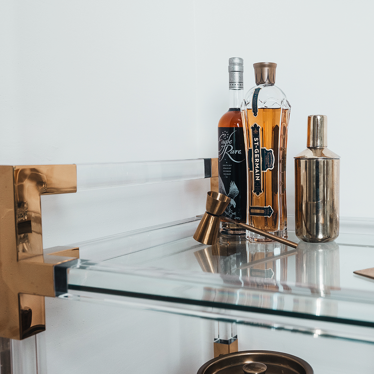 A closeup of the top shelf of an acrylic lucite bar cart featuring gold hardware displaying spirts and various bar accessories.