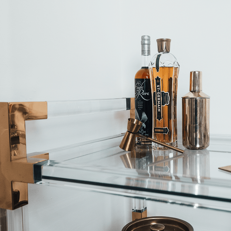 A closeup of the top shelf of an acrylic lucite bar cart featuring gold hardware displaying spirts and various bar accessories.