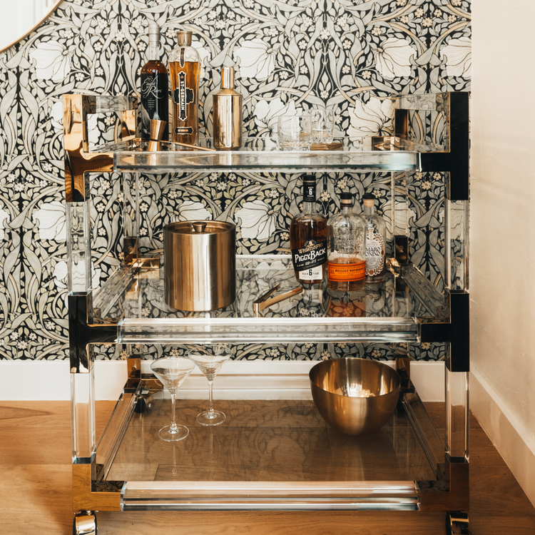 An acrylic lucite bar cart placed in a dining room corner featuring gold hardware, 3 shelves, and wheels for easy movement. The cart is decorated with spirts, glasses, and various bar accessories.