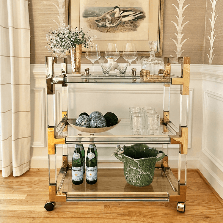 An acrylic lucite bar cart placed in a dining room corner featuring gold hardware, 3 shelves, and wheels for easy movement. The cart is decorated with flowers, spirts, glasses, and various bar accessories.