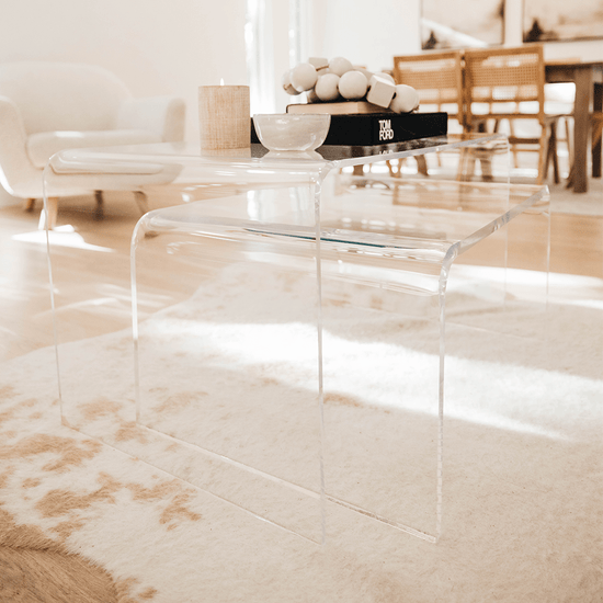 A clear acrylic lucite nesting coffee table set with candles, decor, and books on top placed on a light white and brown cowhide rug in a living room setting.