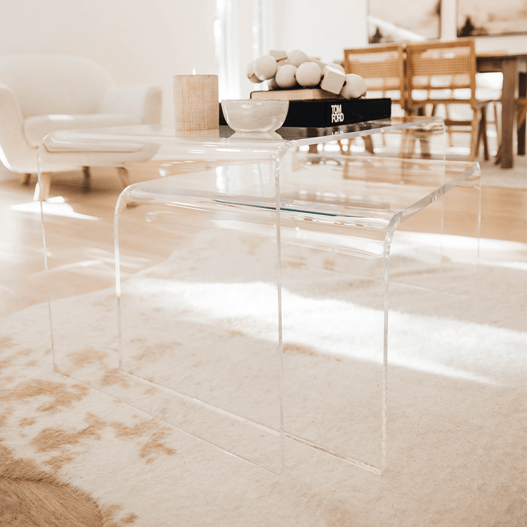 A clear acrylic lucite nesting coffee table set with candles, decor, and books on top placed on a light white and brown cowhide rug in a living room setting.